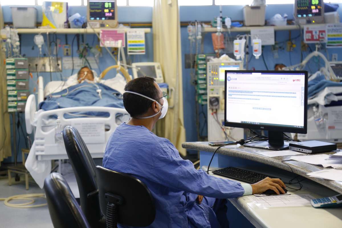 Em uma sala de terapia intensiva, um paciente está deitado no leito com vários equipamentos médicos ao seu redor e um profissional de saúde está sentado usando o computador