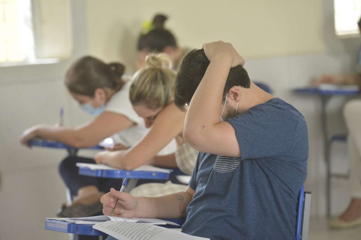 Imagem de quatro candidatos fazendo a prova do concurso Funsaúde. Em primeiro plano, um homem branco, segurando a caneta com a mão direita e a outra sobre a cabeça